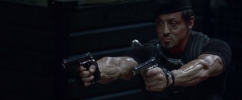 Movie still from “The Expendables” (2010), directed by Sylvester Stallone – A person holding a gun in a room; Close Up shot, Low angle