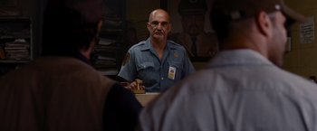 Movie still from “The Expendables” (2010), directed by Sylvester Stallone – An older man in a police uniform is holding a sandwich; Medium shot, Over the shoulder angle