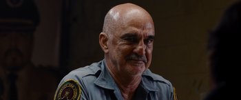 Movie still from “The Expendables” (2010), directed by Sylvester Stallone – An older man in a police uniform looking at the camera; Close Up shot, Over the shoulder angle