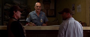Movie still from “The Expendables” (2010), directed by Sylvester Stallone – A man in a police uniform standing behind a counter; Medium shot, Over the shoulder angle