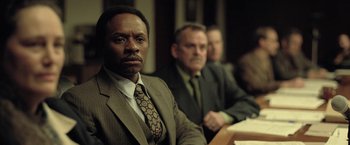 Movie still from “The Fall of the House of Usher” (2023), directed by Mike Flanagan – A man in a suit and tie sitting in front of other men; Close Up shot, Over the shoulder angle