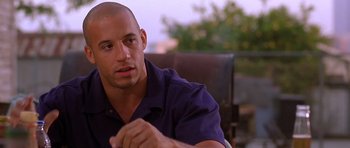 Movie still from “The Fast and the Furious” (2001), directed by Rob Cohen – A man sitting in front of a chair with his hand up to his face; Close Up shot, Over the shoulder angle