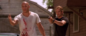 Movie still from “The Fast and the Furious” (2001), directed by Rob Cohen – A man holding a gun next to another man with blood all over; Medium shot, Over the shoulder angle