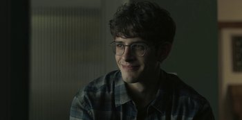 Movie still from “The Girl in the Mirror” (2022), created by Sergio G. Sánchez – A young man wearing glasses and a checkered shirt is smiling; Close Up shot, Over the shoulder angle