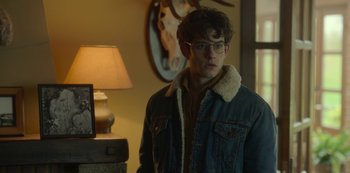 Movie still from “The Girl in the Mirror” (2022), created by Sergio G. Sánchez – A young man wearing glasses and a jean jacket standing next to a lamp; Close Up shot, Over the shoulder angle