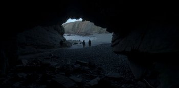 Movie still from “The Girl in the Mirror” (2022), created by Sergio G. Sánchez – Two people are walking on the beach near a cave; Extreme Wide shot, Low angle