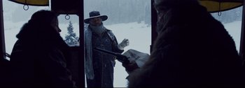Movie still from “The Hateful Eight” (2015), directed by Quentin Tarantino – A man dressed in winter clothing and a hat is standing in the snow looking at a newspaper while another man is holding a gun; Medium shot, Over the shoulder angle