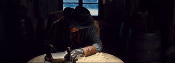 Movie still from “The Hateful Eight” (2015), directed by Quentin Tarantino – A man in a cowboy hat and leather gloves working on a piece of wood; Medium shot, Over the shoulder angle