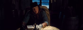 Movie still from “The Hateful Eight” (2015), directed by Quentin Tarantino – A man sitting at a table in a room with a book; Medium shot, Over the shoulder angle