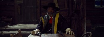 Movie still from “The Hateful Eight” (2015), directed by Quentin Tarantino – An old man sitting at a wooden table wearing a hat; Medium shot, Over the shoulder angle