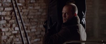 Movie still from “The Italian Job” (2003), directed by F. Gary Gray – A bald man with a beard is holding a guitar case; Close Up shot, Over the shoulder angle