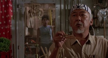 Movie still from “The Karate Kid” (1984), directed by John G. Avildsen – An older man holding chopsticks in front of a woman; Medium shot, Over the shoulder angle