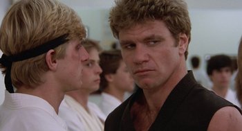 Movie still from “The Karate Kid” (1984), directed by John G. Avildsen – A man in a black shirt is looking at the camera with another man behind him; Close Up shot, Low angle