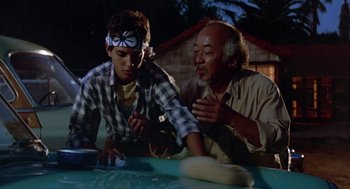 Movie still from “The Karate Kid” (1984), directed by John G. Avildsen – A man and a young boy are making something out of dough; Medium shot, Over the shoulder angle