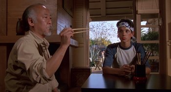 Movie still from “The Karate Kid” (1984), directed by John G. Avildsen – Two people sitting at a table with chopsticks in their hands; Medium shot, Over the shoulder angle