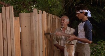 Movie still from “The Karate Kid” (1984), directed by John G. Avildsen – A man and a boy standing in front of a wooden fence; Medium shot, Over the shoulder angle