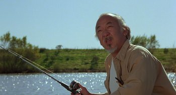 Movie still from “The Karate Kid” (1984), directed by John G. Avildsen – An older man holding a fishing pole while standing in front of a body of water; Medium shot, Low angle