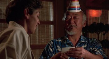 Movie still from “The Karate Kid” (1984), directed by John G. Avildsen – A man wearing a party hat is giving a birthday card; Close Up shot, Over the shoulder angle