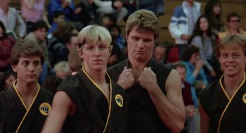 Movie still from “The Karate Kid” (1984), directed by John G. Avildsen – A man standing next to a young man in a black shirt; Close Up shot, Low angle