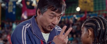 Movie still from “The Karate Kid” (2010), directed by Harald Zwart – An asian man in a blue and white jacket is holding his hand up to his face; Close Up shot, Over the shoulder angle