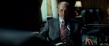 Movie still from “The Kingdom” (2007), directed by Peter Berg – An older man wearing a suit and tie sitting at a table; Medium shot, Over the shoulder angle
