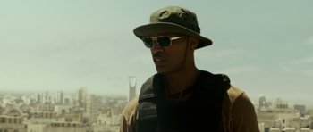 Movie still from “The Kingdom” (2007), directed by Peter Berg – A man wearing sunglasses and a hat is standing in front of a city skyline; Close Up shot, Low angle