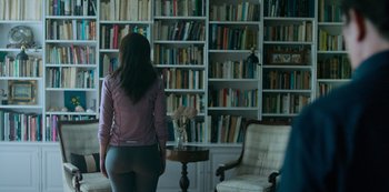 Movie still from “The Longest Night” (2022), directed by Moisés Ramos – A woman standing in front of bookshelves in a living room; Medium shot, Over the shoulder angle