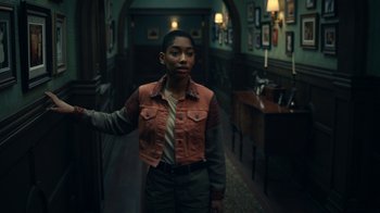 Movie still from “The Midnight Club” (2022), created by Mike Flanagan – A person standing in a hallway with a lamp on; Medium shot, Over the shoulder angle