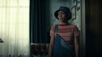 Movie still from “The Midnight Club” (2022), created by Mike Flanagan – A woman wearing a hat and overalls in a living room; Medium shot, Low angle