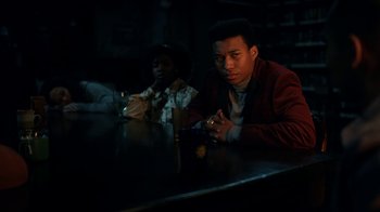 Movie still from “The Midnight Club” (2022), created by Mike Flanagan – A man sitting at a table with a drink in front of him; Medium shot, Over the shoulder angle