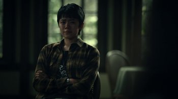 Movie still from “The Midnight Club” (2022), created by Mike Flanagan – A person sitting in a chair with his arms crossed; Medium shot, Over the shoulder angle