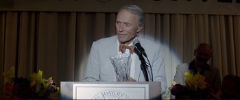 Movie still from “The Mule” (2018), directed by Clint Eastwood – An older man in a suit and bow tie holding a glass trophy; Medium shot, Over the shoulder angle