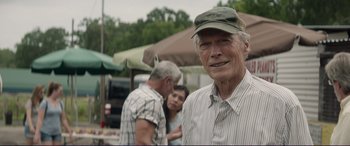 Movie still from “The Mule” (2018), directed by Clint Eastwood – An older man wearing a hat standing in front of other people; Close Up shot, Over the shoulder angle