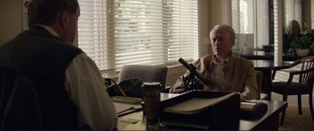 Movie still from “The Mule” (2018), directed by Clint Eastwood – An older man sitting at a table with a microphone; Medium shot, Over the shoulder angle