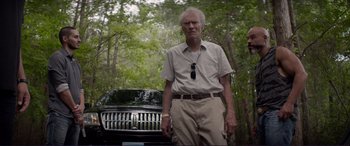 Movie still from “The Mule” (2018), directed by Clint Eastwood – An older man standing in front of a car in a wooded area; Medium shot, Low angle