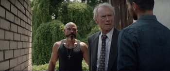Movie still from “The Mule” (2018), directed by Clint Eastwood – A man in a black shirt and a man in a suit and tie standing next to each other in front of trees; Medium shot, Over the shoulder angle