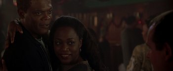 Movie still from “The Negotiator” (1998), directed by F. Gary Gray – A woman smiles at the camera with a man in the background; Close Up shot, Over the shoulder angle