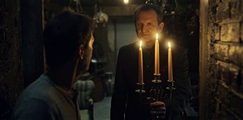Movie still from “Guillermo del Toro's Cabinet of Curiosities” (2022), directed by Keith Thomas – A man in a suit holding two lit candles in front of him; Medium shot, Over the shoulder angle