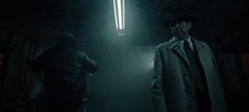 Movie still from “Guillermo del Toro's Cabinet of Curiosities” (2022), directed by Keith Thomas – A man in a trench coat and hat standing in a dimly - lit room; Medium shot, Low angle