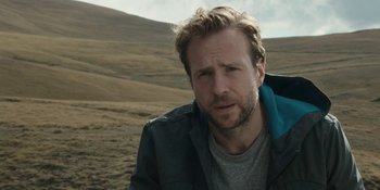 Movie still from “The Ritual” (2017), directed by David Bruckner – A person wearing a jacket near a field; Close Up shot, Low angle