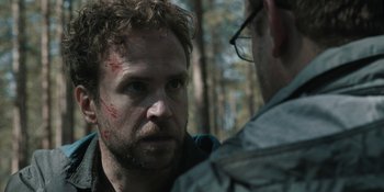 Movie still from “The Ritual” (2017), directed by David Bruckner – A man with a scar on his face talking to another man in the woods; Close Up shot, Over the shoulder angle