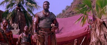Movie still from “The Scorpion King” (2002), directed by Chuck Russell – A man in a leather outfit standing next to palm trees; Medium shot, Low angle