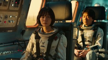 Movie still from “The Silent Sea” (2021), created by Choi Hang-Yong – A woman sitting in the back of an airplane wearing a space suit; Medium shot, Over the shoulder angle