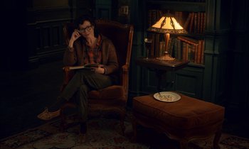 Movie still from “Guillermo del Toro's Cabinet of Curiosities” (2022), directed by Keith Thomas – A woman sitting in a chair reading a book in front of a lamp; Wide shot, High angle