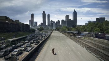 Movie still from “The Walking Dead” (2010), directed by Daniel Sackheim – A person standing on the side of a road in front of a city skyline; Extreme Wide shot, High angle