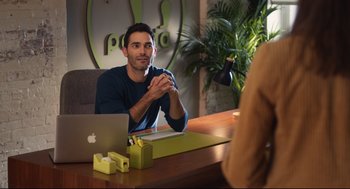 Movie still from “Can You Keep a Secret?” (2019), directed by Elise Durán – A man sitting in front of a laptop computer on top of a wooden table; Medium shot, Over the shoulder angle