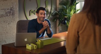 Movie still from “Can You Keep a Secret?” (2019), directed by Elise Durán – A man sitting in front of a laptop computer on top of a wooden table; Medium shot, Over the shoulder angle