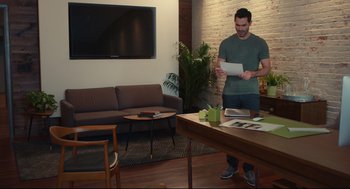 Movie still from “Can You Keep a Secret?” (2019), directed by Elise Durán – A man standing in a living room holding papers; Wide shot, High angle