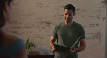 Movie still from “Can You Keep a Secret?” (2019), directed by Elise Durán – A man standing in front of a table holding a book; Medium shot, Over the shoulder angle
