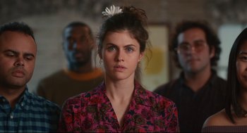 Movie still from “Can You Keep a Secret?” (2019), directed by Elise Durán – A woman with a flower in her hair stands in front of a group of people; Close Up shot, Over the shoulder angle